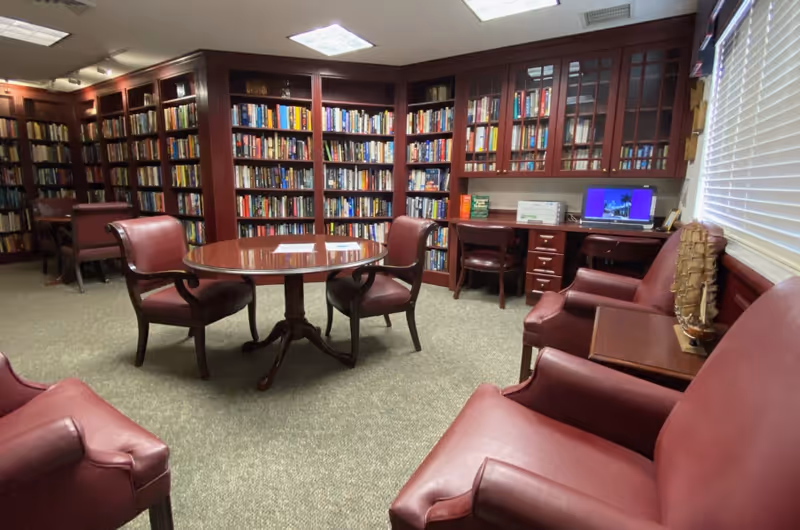 A cozy library/reading room with wood bookshelves lining the walls, a round table and red leather chairs.