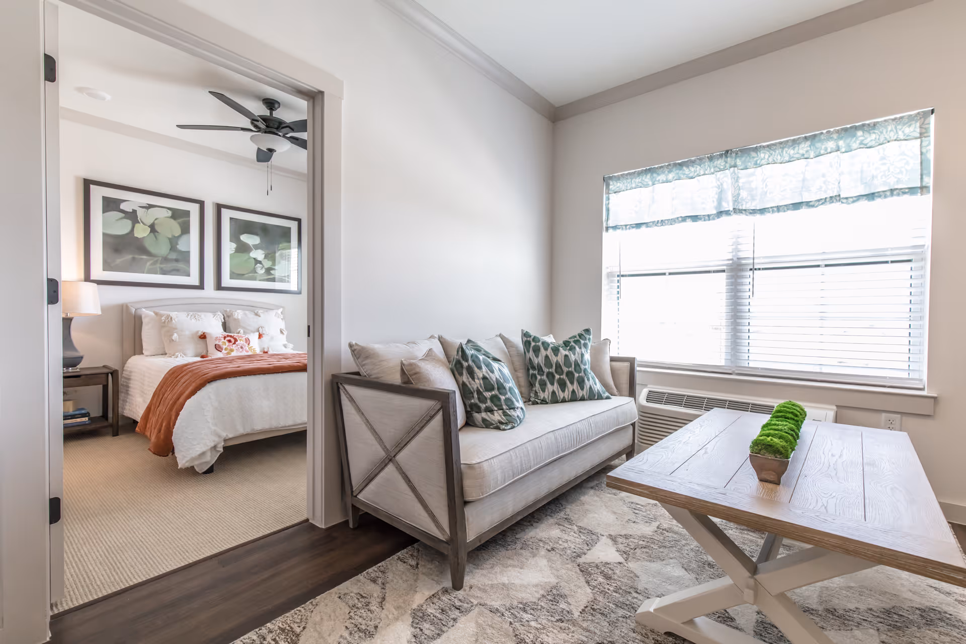 A bright living room with a beige sofa adorned with patterned throw pillows, a wooden coffee table with a small green plant centerpiece, and a large window with blinds and a valance. Through an open doorway, a bedroom is visible featuring a bed with white bedding, a rust-colored throw blanket, two framed botanical prints above the headboard, a nightstand with a lamp, and a ceiling fan.