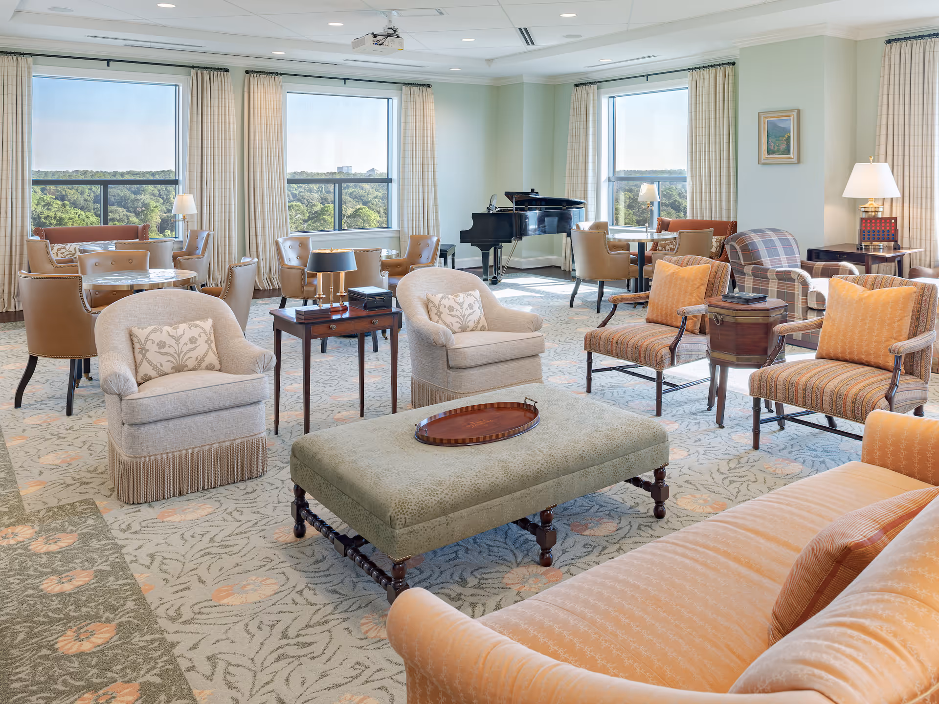 A spacious, well-lit living room with large windows showing a green outdoor view. The room features multiple seating areas including beige armchairs, striped chairs with orange cushions, a large ottoman with a tray, and a peach-colored sofa. There are several small tables with lamps and decorative items, and a black grand piano is positioned near the windows.