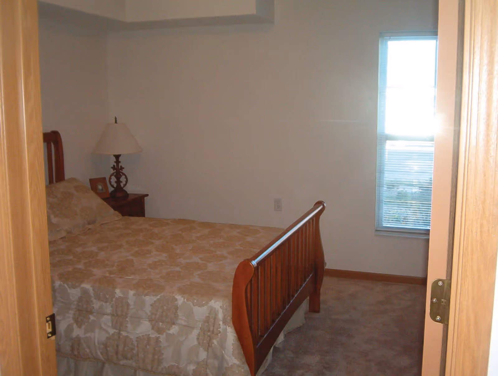 A bedroom with a wooden bed frame and a beige patterned bedspread. There is a wooden nightstand with a decorative lamp and a small framed photo. A window with blinds is on the right wall, letting in sunlight.
