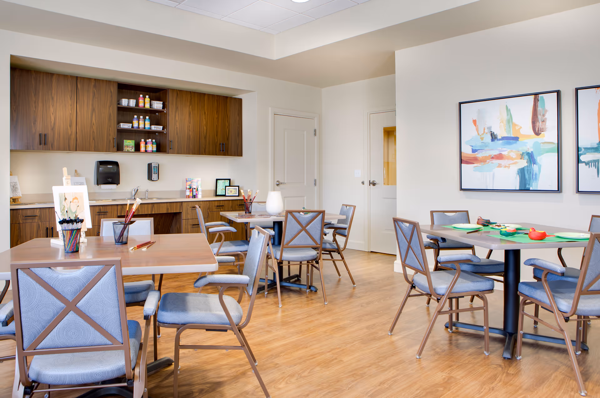 A bright and clean activity room with wooden floors, several tables and chairs, and a kitchenette area with wooden cabinets. The tables have art supplies like paintbrushes, colored pencils, and paper, indicating a space for creative activities. Two abstract paintings hang on the wall.