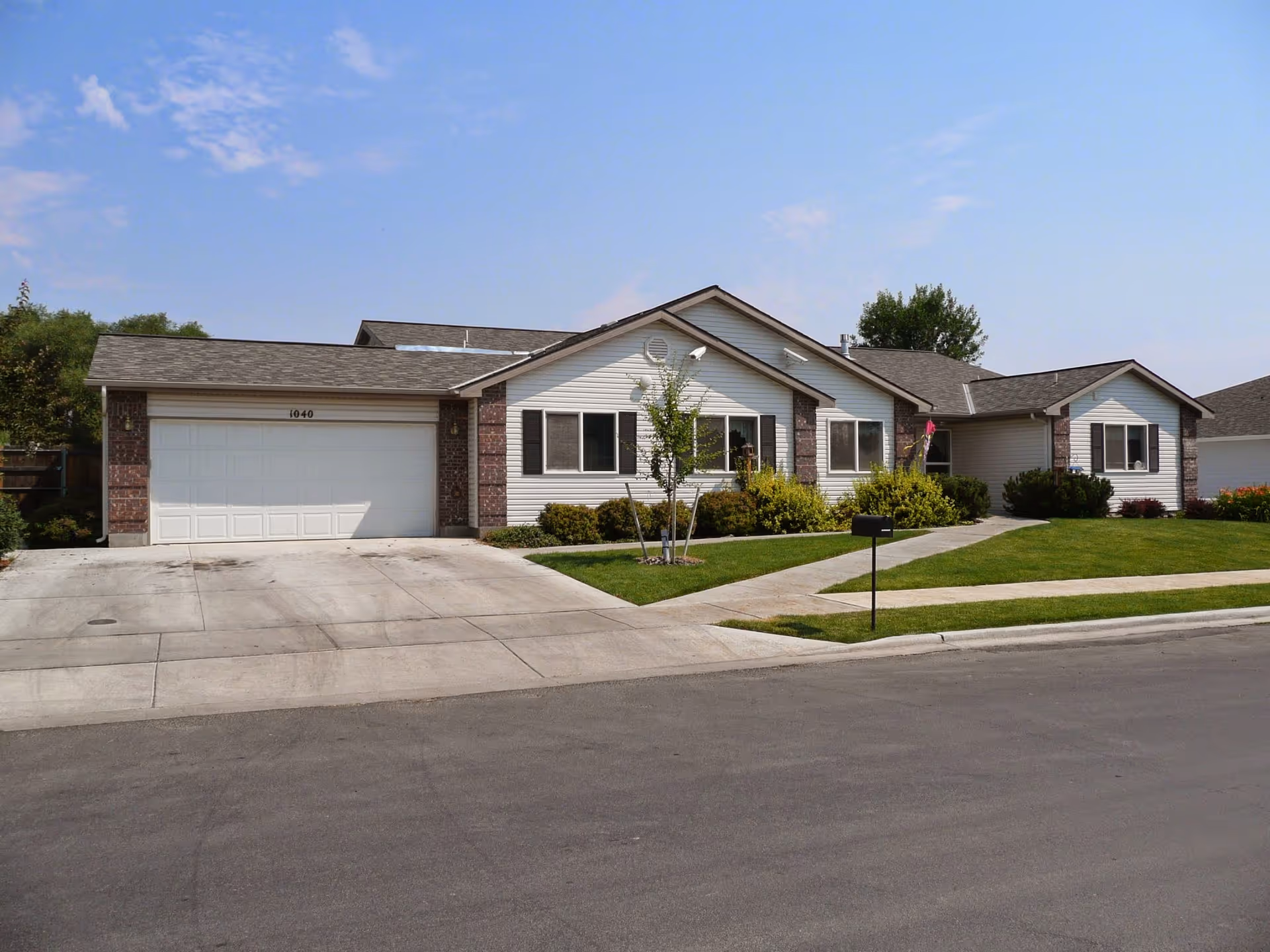 Single-story residential building with a two-car garage, white siding, and brick accents. The house is surrounded by a well-maintained lawn, small trees, and shrubs. The driveway and sidewalk are concrete, and the sky is clear with a few clouds.