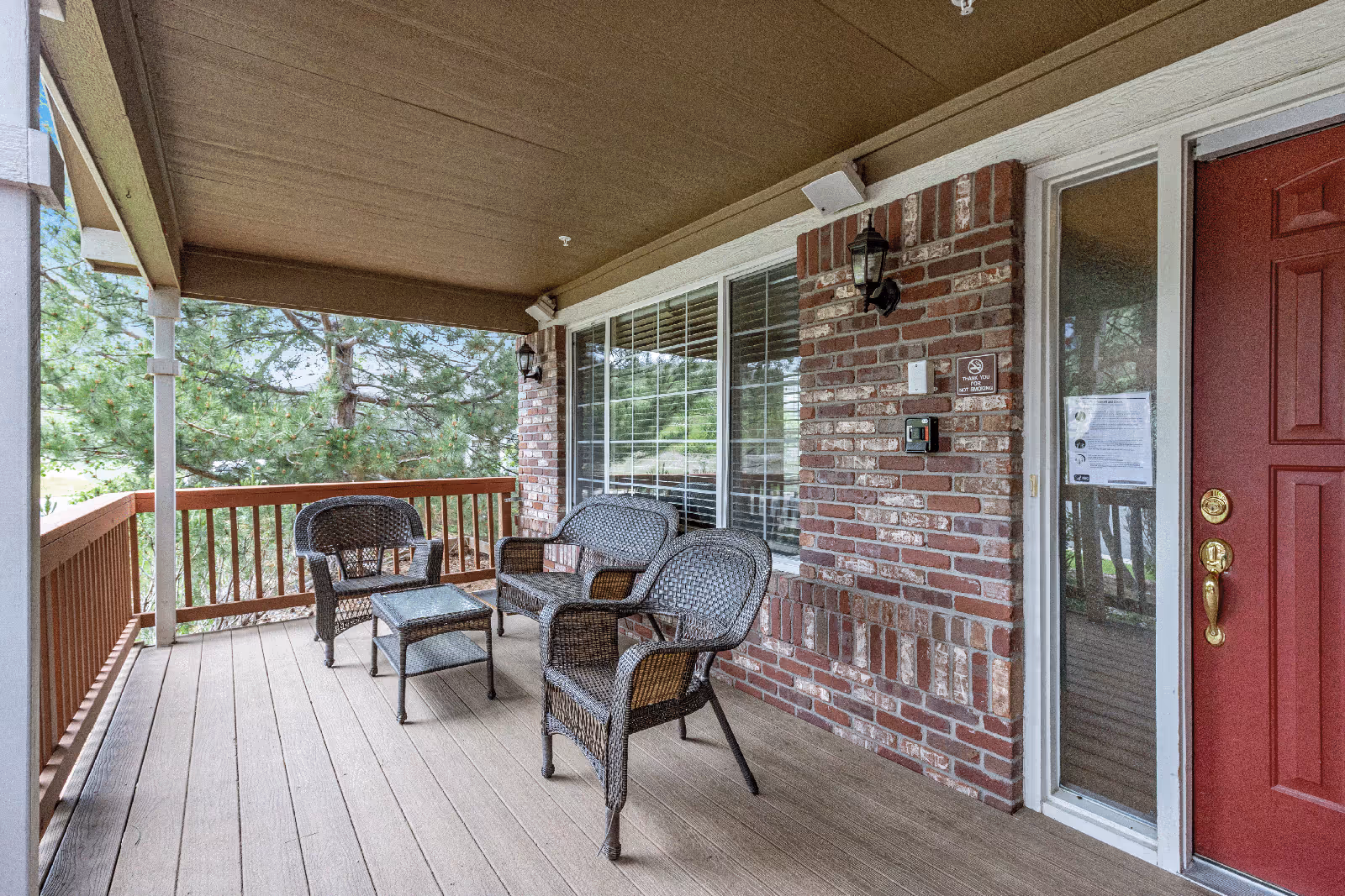 Covered porch area with a wooden floor and railing, featuring three wicker chairs and a small glass-top table. The porch is attached to a brick wall with a large window and a red door with a brass handle. There are two wall-mounted lantern-style lights and a no smoking sign on the brick wall. Trees and greenery are visible beyond the railing.