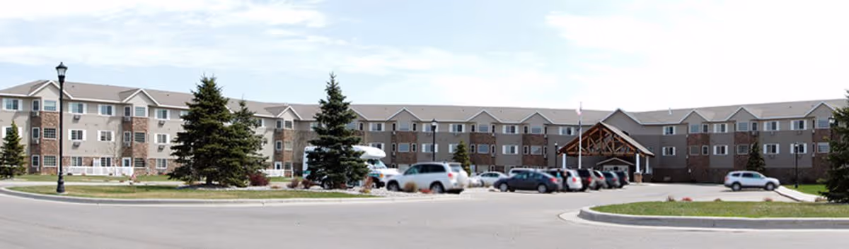 Exterior view of a large three-story senior living facility with a parking lot in front, several cars parked, and a covered entrance. The building has a combination of brick and siding with multiple windows and some evergreen trees in the landscaped area.