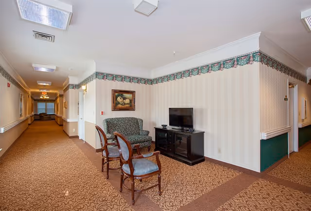 Hallway seating nook in a senior living facility with upholstered chairs, a loveseat, and a TV on a stand.