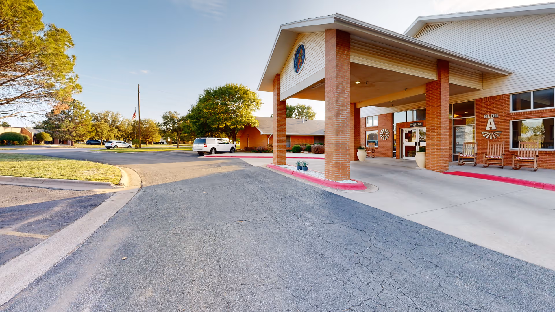 Covered main entrance of an assisted-living building with brick columns, a drive-up canopy, parked cars, and rocking chairs by the doorway.