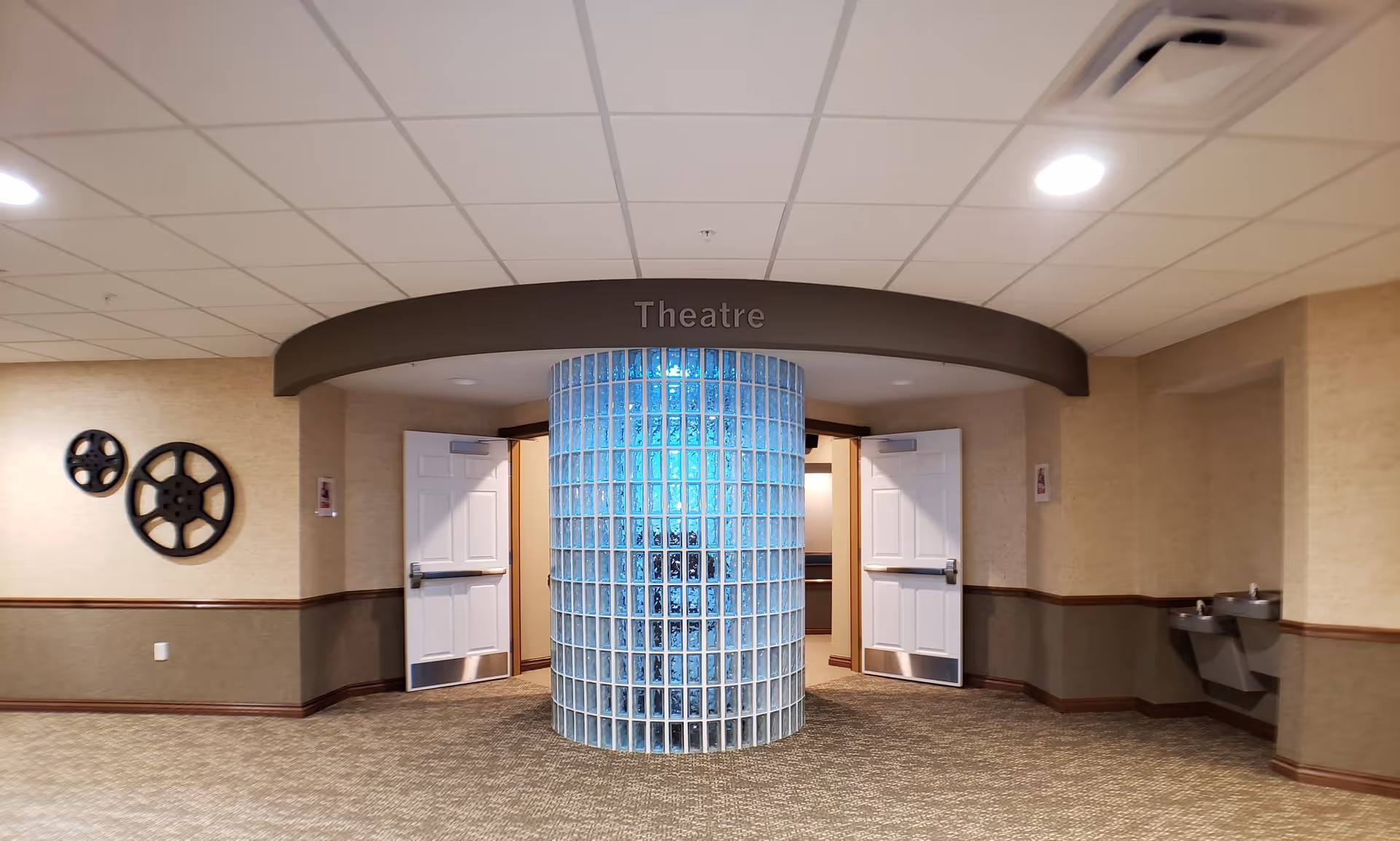 Interior hallway with beige walls and carpeted floor leading to a theatre entrance. The entrance features a curved glass block wall in the center and two white double doors on either side. Above the entrance is a curved sign that reads 'Theatre'. On the left wall, there are two decorative film reels mounted. On the right side, there are two water fountains attached to the wall.
