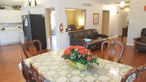 Open-plan dining and living area featuring a patterned dining table with a floral centerpiece, leather sofas, and a kitchen in the background.