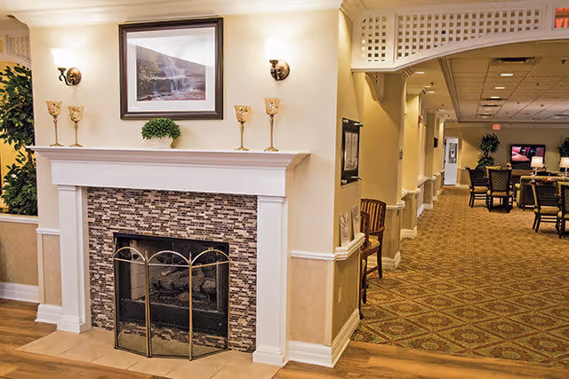 Interior view of a senior living facility featuring a fireplace with a decorative screen, a framed picture above it, and candle holders on the mantel. To the right, a hallway leads to a common area with chairs and tables, illuminated by ceiling lights.