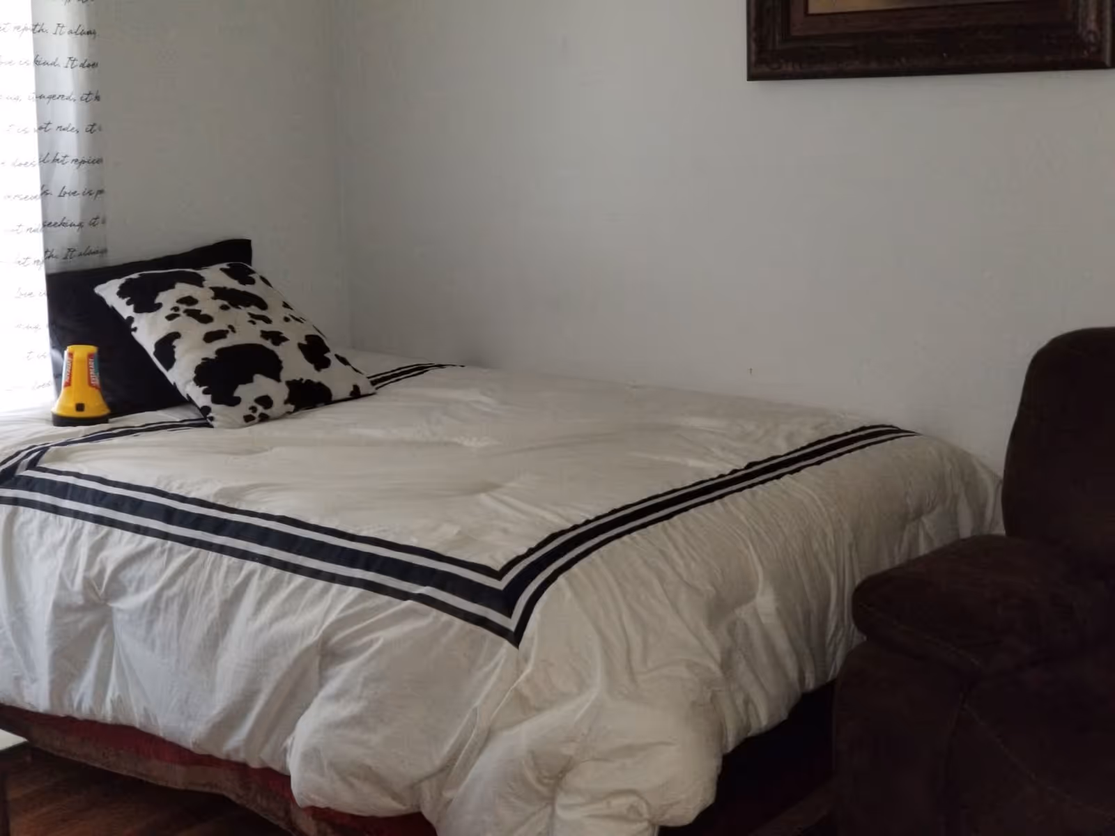 A bedroom with a bed covered in a white comforter with black stripes. There are two pillows on the bed, one black and one with a black and white cow print. A yellow flashlight is placed near the pillows. A brown armchair is partially visible on the right side of the image. The walls are plain white with a framed picture hanging above the bed.