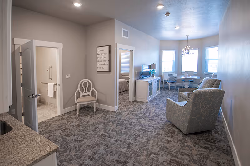 A spacious assisted living apartment interior featuring a living area with two patterned armchairs, a TV on a white cabinet, a round dining table with chairs near three windows, and an open doorway leading to a bedroom and a bathroom. The room has light-colored walls, recessed ceiling lights, and carpeted flooring.
