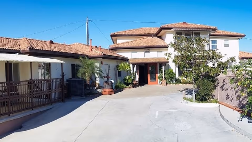 Exterior view of King's Care Assisted Living LLC building with a driveway leading to the entrance. The building has a tiled roof, white walls, and some greenery including trees and plants near the entrance.