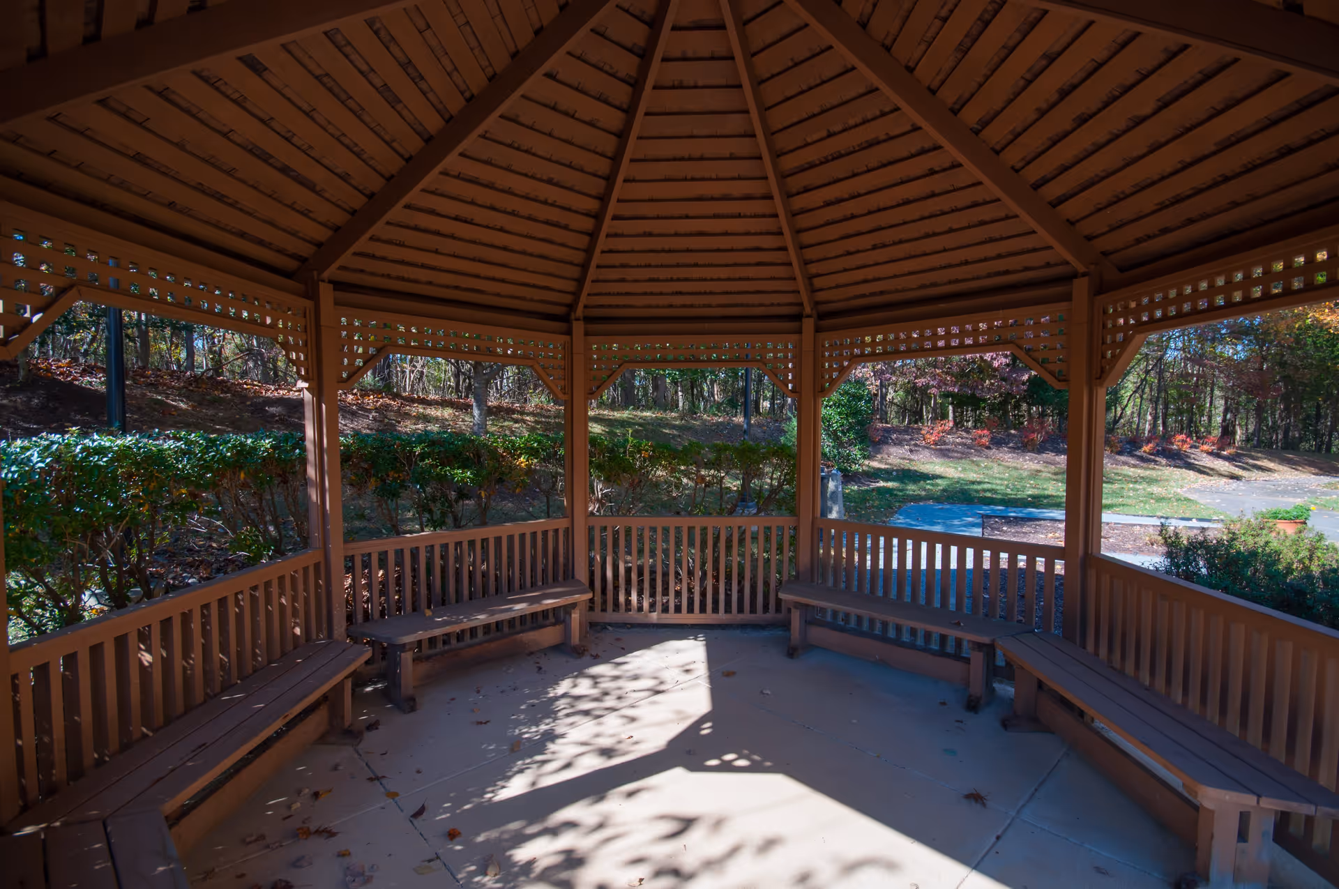 A wooden gazebo with a peaked roof and built-in benches along the perimeter, situated outdoors surrounded by trees and greenery under natural daylight.