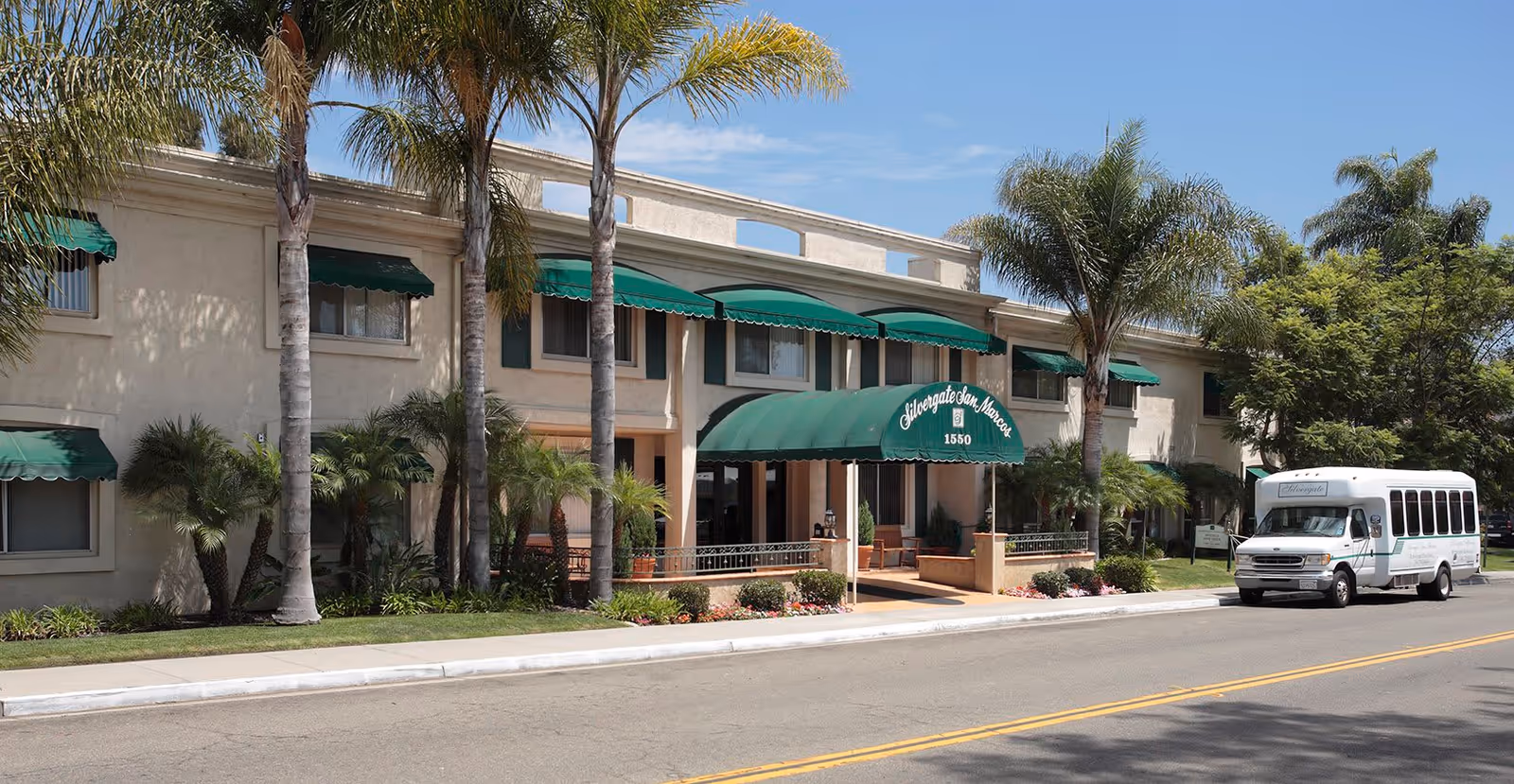 Exterior view of Silvergate San Marcos senior living facility showing a two-story building with green awnings over the windows and entrance. Palm trees and other landscaping are visible in front of the building. A white shuttle bus is parked on the street to the right.