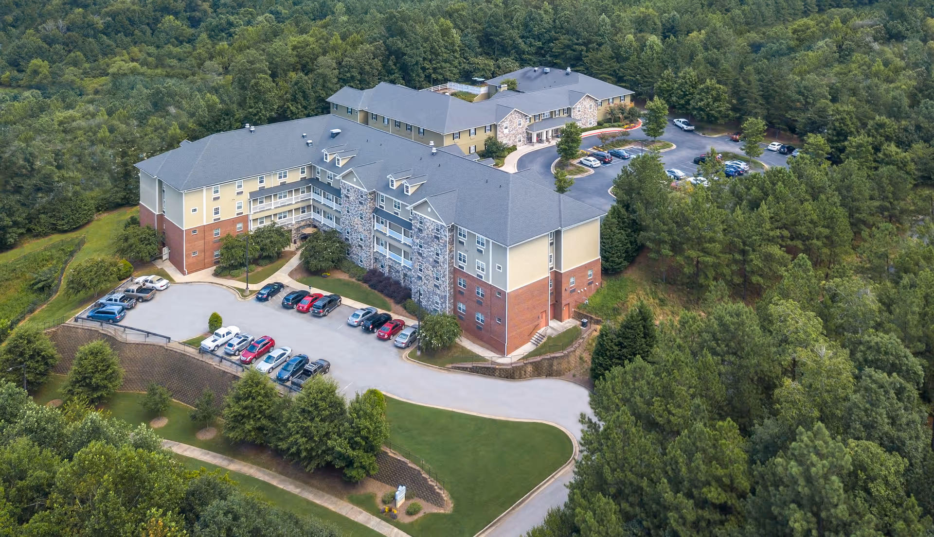 Aerial view of a multi-story senior living building with parking lots, landscaped grounds, and surrounding trees.