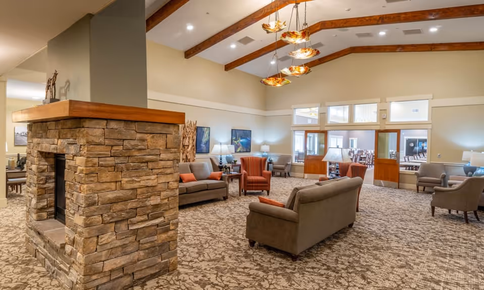 A spacious living room area with beige walls and carpeted floor featuring a stone fireplace with a wooden mantle in the foreground. The room has multiple seating options including sofas and armchairs with orange accent pillows. Wooden beams run across the ceiling with decorative hanging lights. Large windows and glass doors lead to an adjacent dining area.