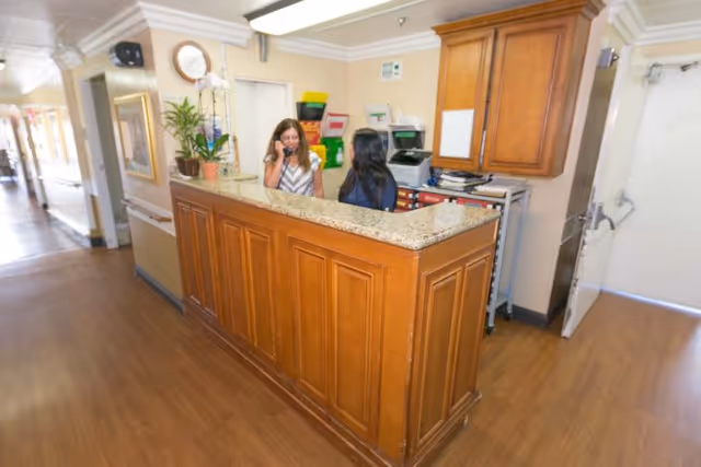 Reception area in a nursing center with a wooden counter and two women behind it, one talking on the phone. The area has wooden flooring, a clock on the wall, and some plants and office supplies on the counter.
