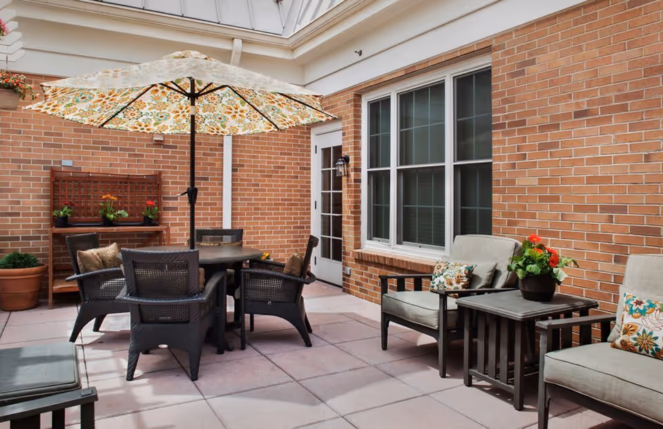 Outdoor patio area with a round table surrounded by four wicker chairs, a large floral-patterned umbrella providing shade, cushioned armchairs with floral pillows, a small side table with a potted plant, and a brick wall with windows and a door in the background.