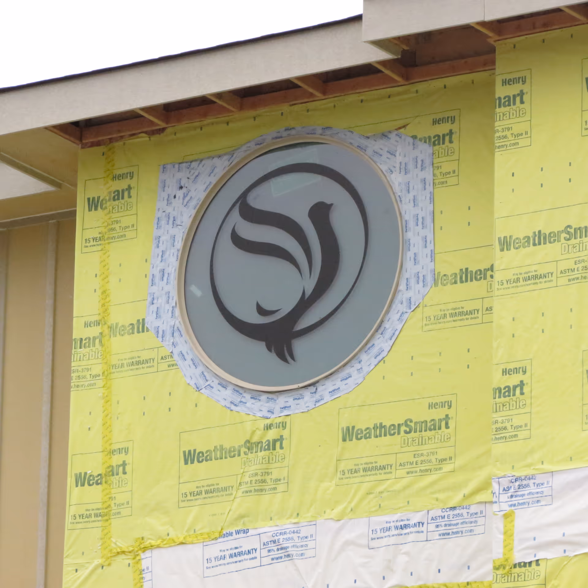 Circular frosted window with a black emblem set in a building facade covered in yellow WeatherSmart house wrap during construction.
