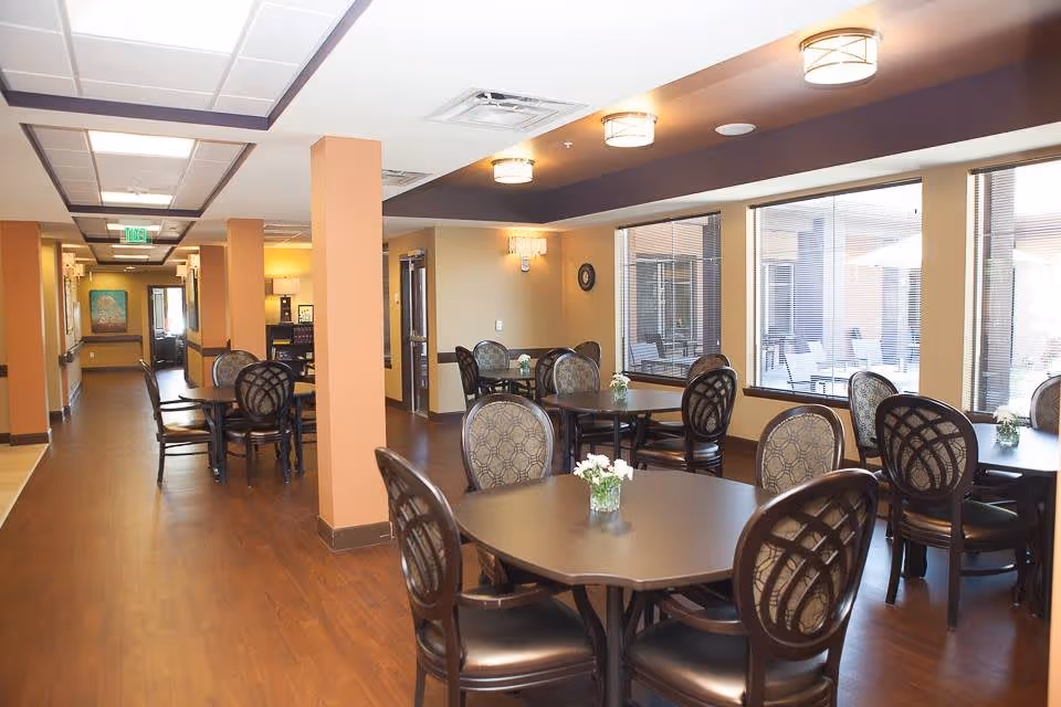 Interior view of a dining area in a senior living facility with multiple dark wood tables and cushioned chairs arranged neatly. Small flower arrangements are placed on each table. Large windows on one side let in natural light and show an outdoor patio area with umbrellas and chairs. The room has warm-toned walls, wooden flooring, and ceiling lights.