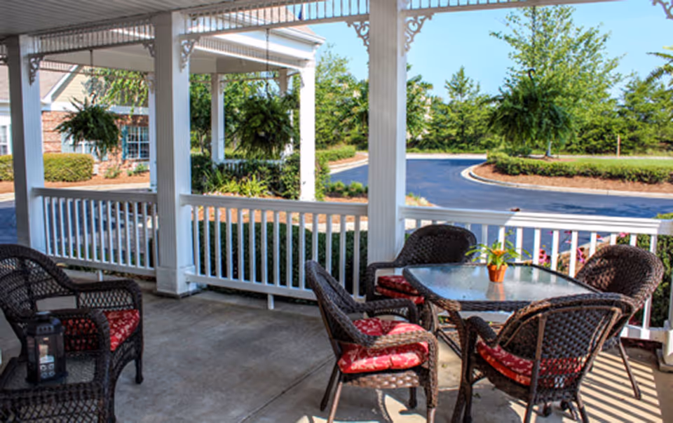 Covered outdoor patio with wicker chairs and a glass-top table overlooking a driveway and landscaped grounds.