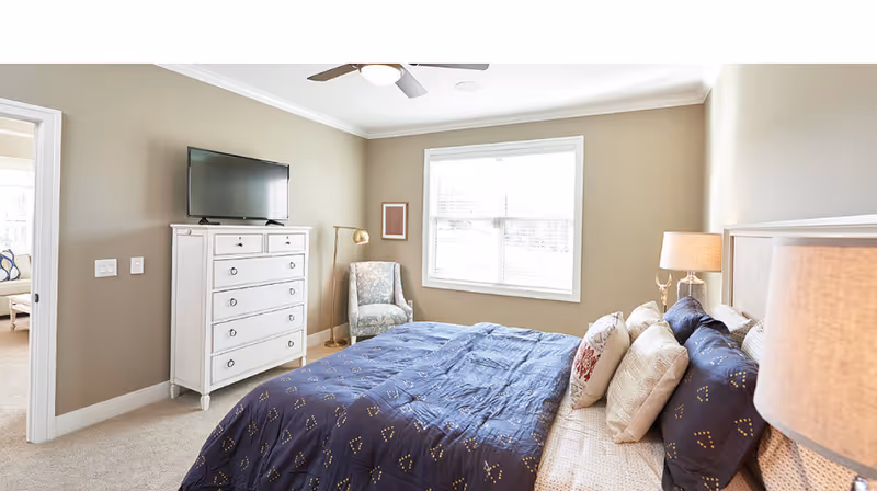 A bright bedroom with a large window letting in natural light. The room features a bed with a blue patterned comforter and multiple pillows, a white chest of drawers with a flat-screen TV on top, a patterned armchair next to a gold floor lamp, and a bedside table with a lamp. The walls are painted beige and the carpet is light-colored.