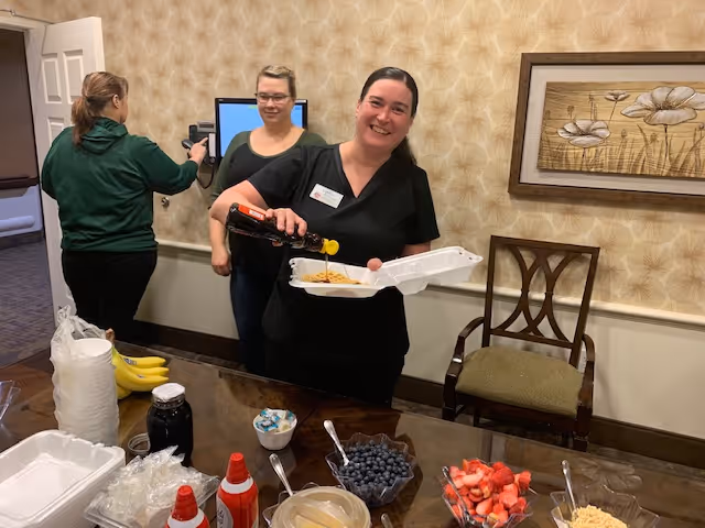 A smiling staff member pours syrup into a takeout container at a buffet table with bowls of fruit and toppings in a dining room while two other women stand nearby.