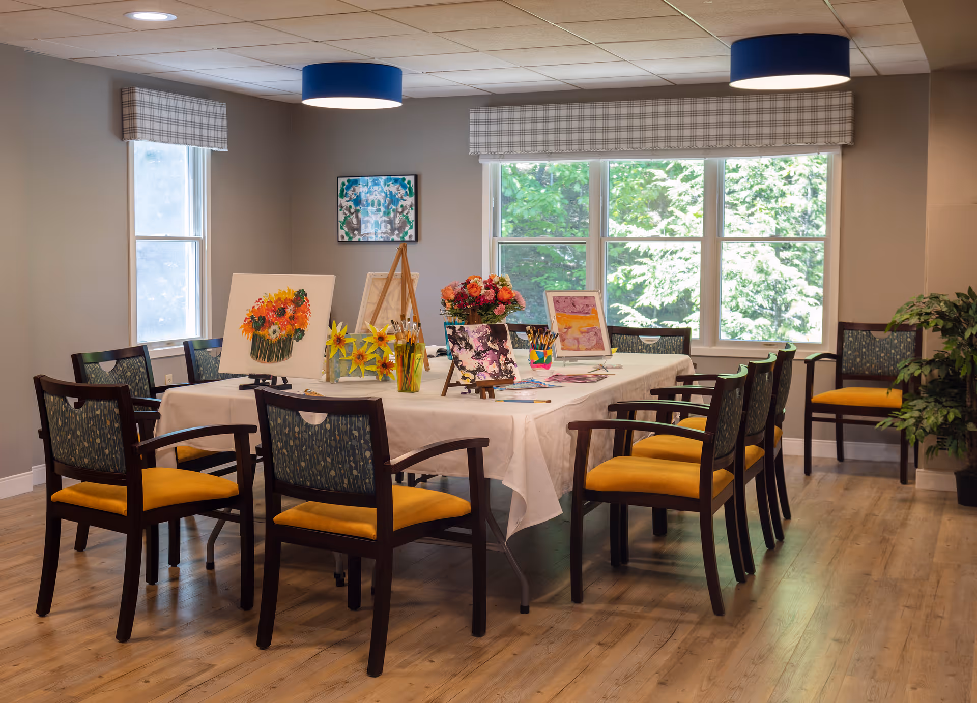A bright assisted-living activity room with a table covered in painting supplies and surrounded by chairs in front of large windows.