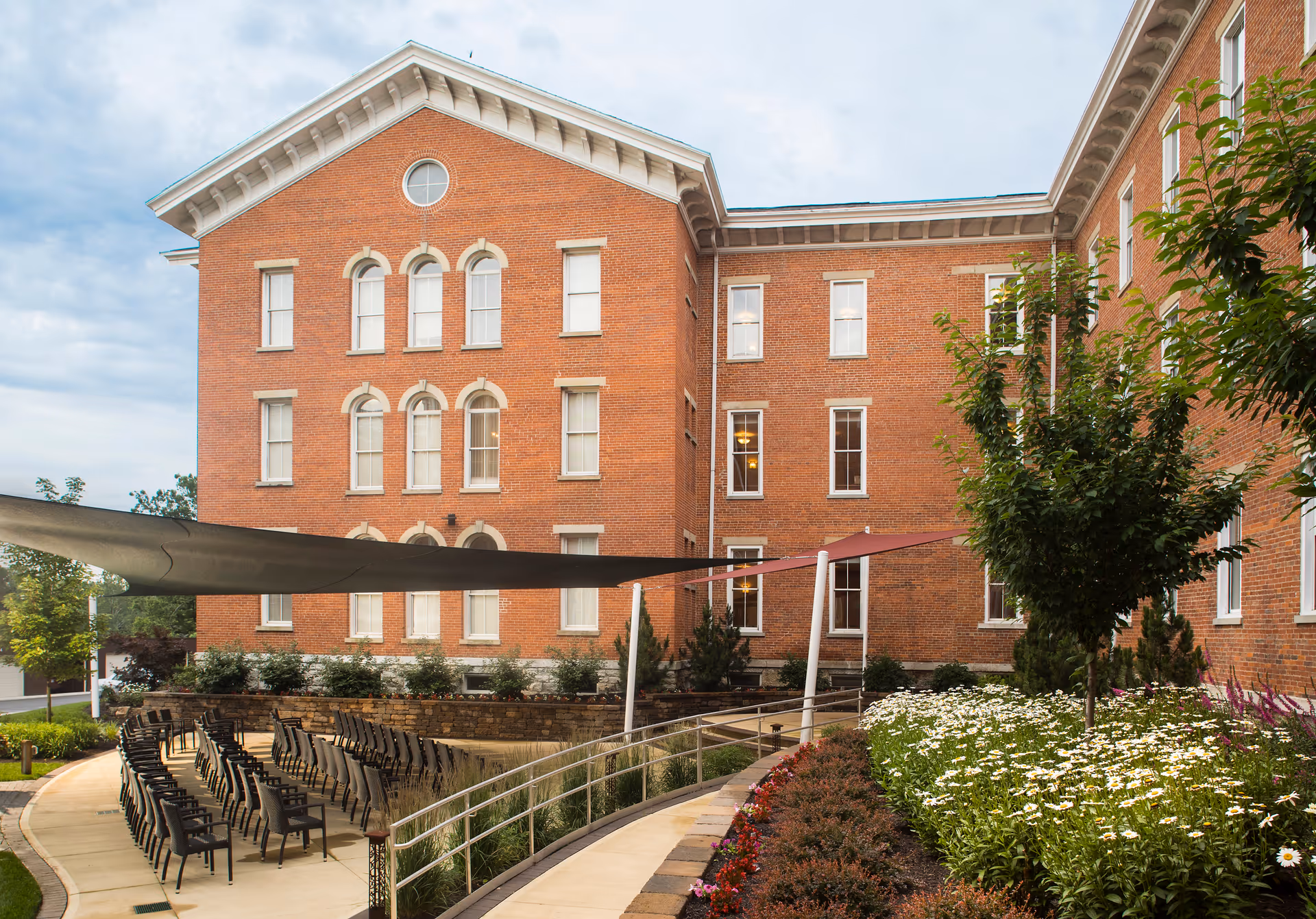 Outdoor courtyard area of a brick retirement community building with rows of black chairs arranged on a paved surface under shade sails, surrounded by landscaped flower beds and trees.