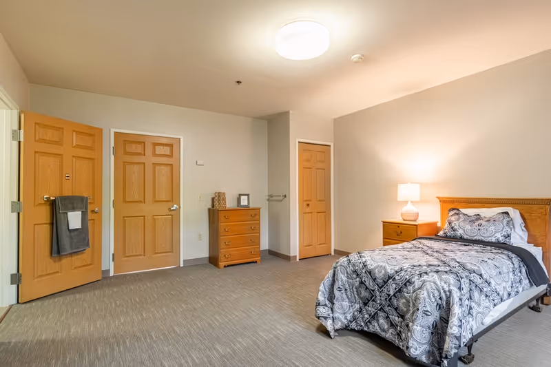 A simple bedroom in a senior living facility with a single bed covered in a patterned gray and white comforter, a wooden nightstand with a lamp, a wooden dresser, and three wooden doors. The room has beige walls and carpeted flooring.