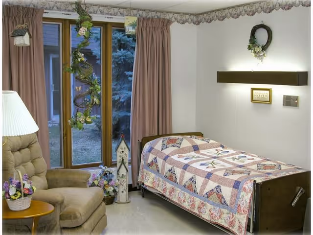 A cozy bedroom in a senior living facility featuring a single bed with a colorful patchwork quilt, a cushioned armchair next to a small wooden side table with a flower basket, large windows with pink curtains, and decorative floral accents on the walls and hanging near the window.