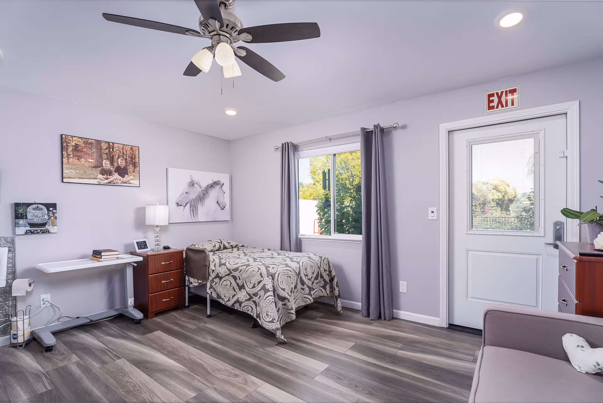 A neatly arranged bedroom in an elder care facility with a single bed covered in a patterned bedspread, a wooden nightstand with a lamp and books, a window with gray curtains, a ceiling fan with lights, a door with an exit sign above it, and wall art including a photo of two children and a drawing of horses.