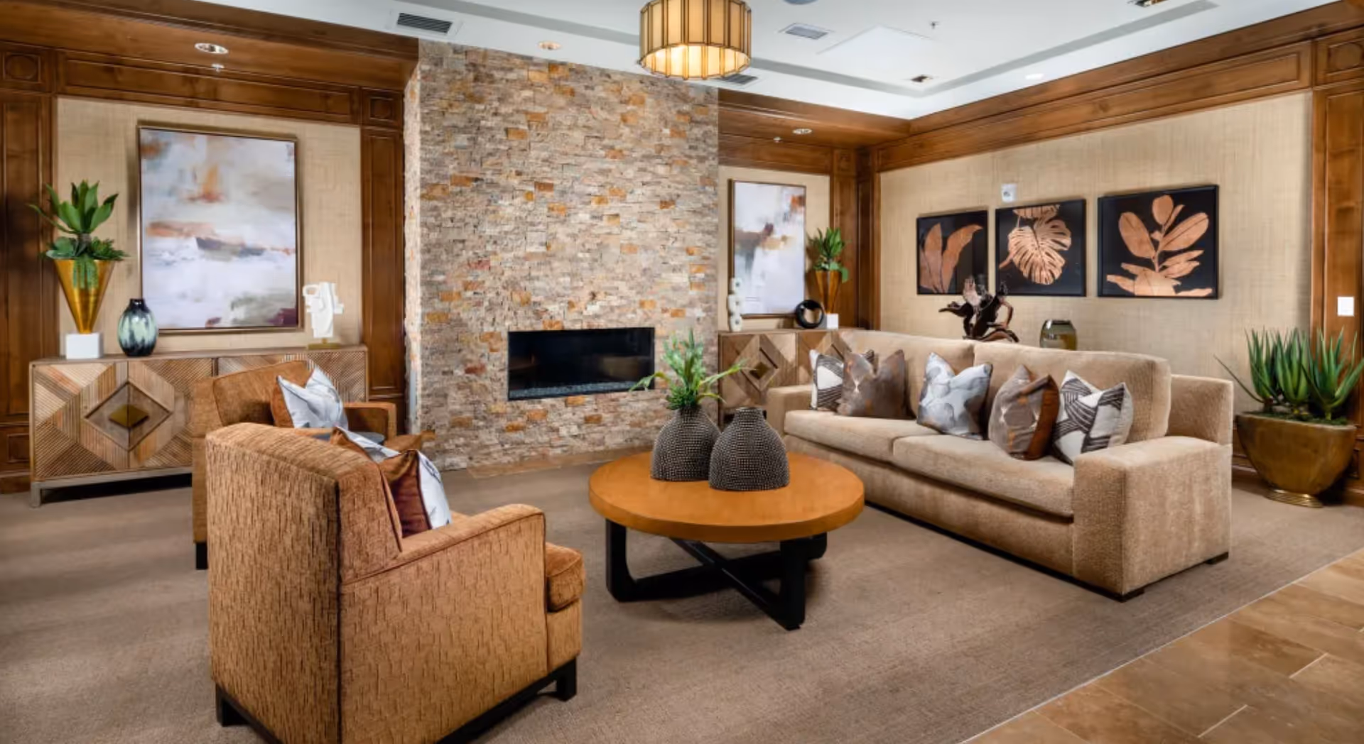 A cozy living room with a beige sofa and two armchairs arranged around a round wooden coffee table. The room features a stone fireplace in the center, wooden paneling on the walls, decorative plants, and framed artwork with leaf motifs. The floor is carpeted and the lighting includes a ceiling fixture.