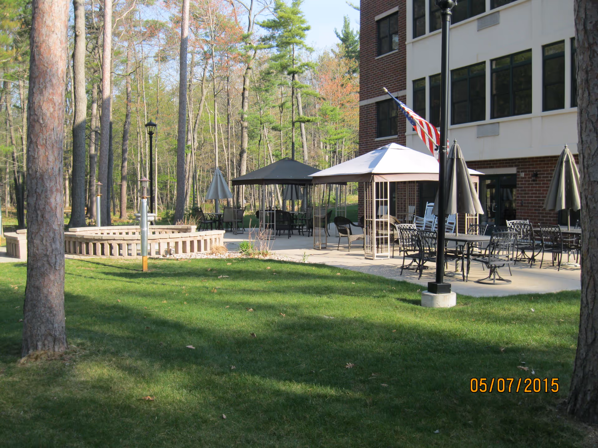 Outdoor patio area at Point Manor Assisted Living with tables, chairs, umbrellas, a gazebo, an American flag on a lamp post, and a grassy lawn surrounded by trees.