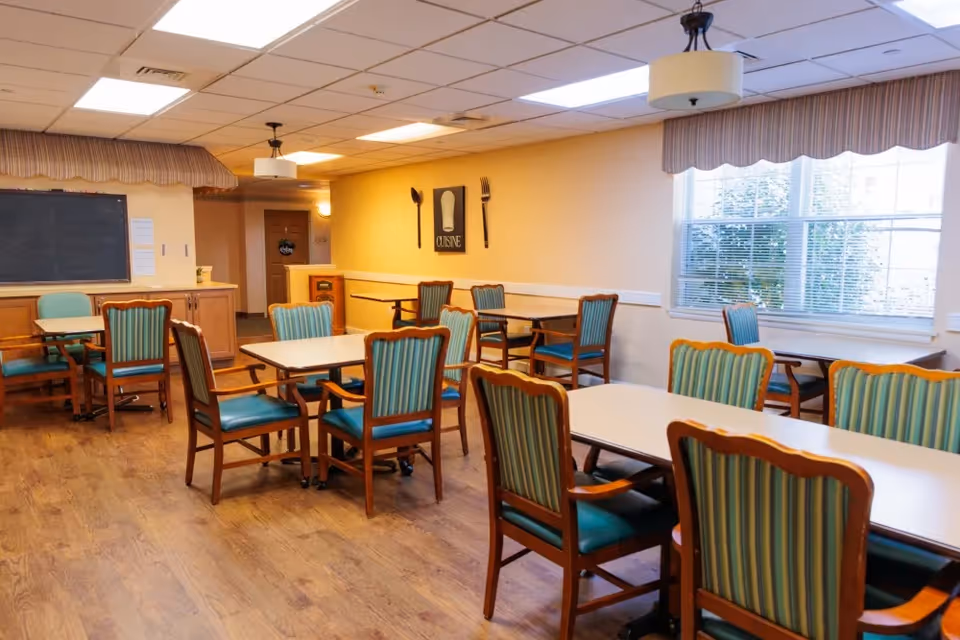 A dining room with several tables and wooden chairs with teal cushions and striped backs. The room has wood flooring, beige walls, and large windows with blinds and valances. There are two ceiling lights and a wall decoration featuring a fork, spoon, and a chef's hat with the word 'CUISINE'.