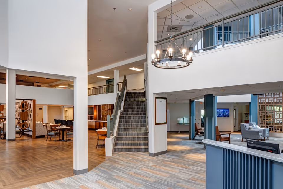 Interior view of a senior living facility lobby with a staircase in the center, seating areas with chairs and tables on the left and right, modern lighting fixtures, and a reception desk in the foreground.