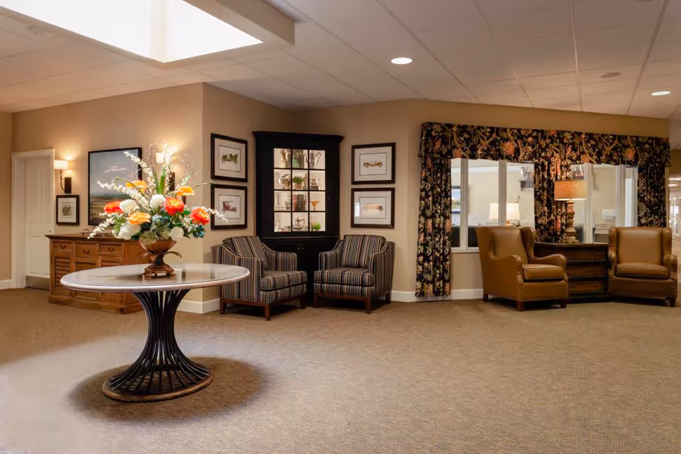 A cozy seating area in a memory care community with a round table holding a large floral arrangement in the center. The room features two striped armchairs next to a black display cabinet with glass doors, framed artwork on the walls, and two brown leather armchairs beside a wooden side table with a lamp. The window is adorned with floral-patterned curtains.
