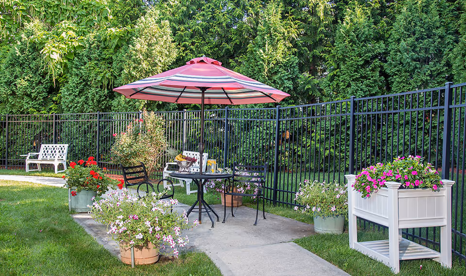 Outdoor patio area with a round table and two black metal chairs under a red and white striped umbrella. The patio is surrounded by various flower pots with blooming flowers and greenery, with a black metal fence and tall green trees in the background. There are also white benches along the fence.