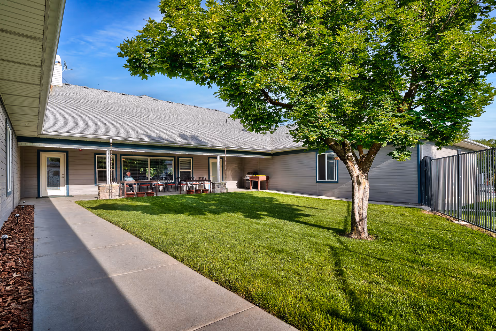 A sunny outdoor courtyard area at Meadow View Senior Living featuring a well-maintained green lawn, a large leafy tree, a concrete walkway, and a covered patio with tables and chairs. A person is seated at one of the tables under the patio. The building exterior is light gray with white trim and a gray shingled roof.