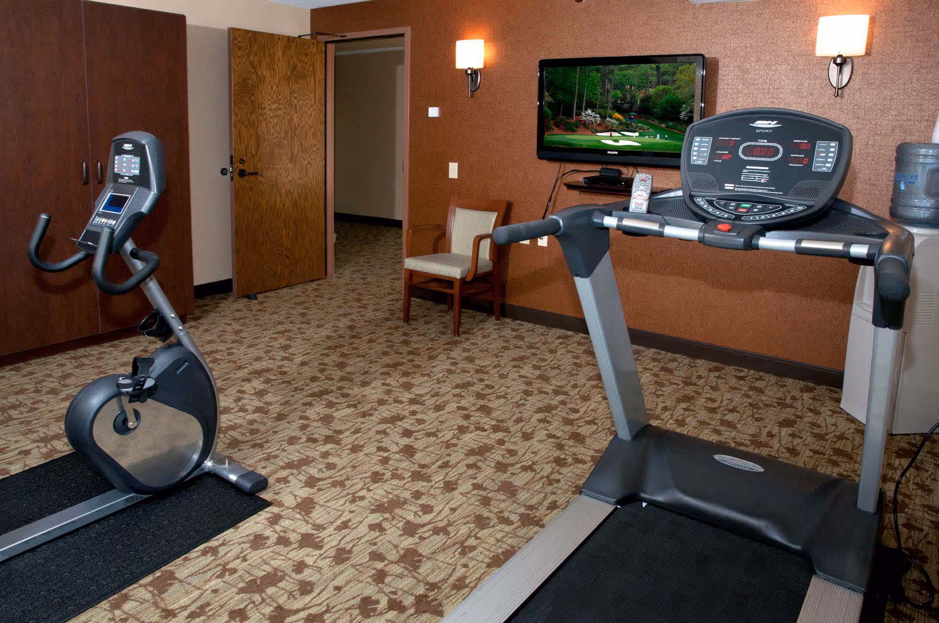 Indoor exercise room with a treadmill and stationary bike on patterned carpet. A wooden chair is placed against a brown wall with two wall-mounted lights. A flat-screen TV is mounted on the wall showing a golf course scene. A water cooler is visible in the corner.