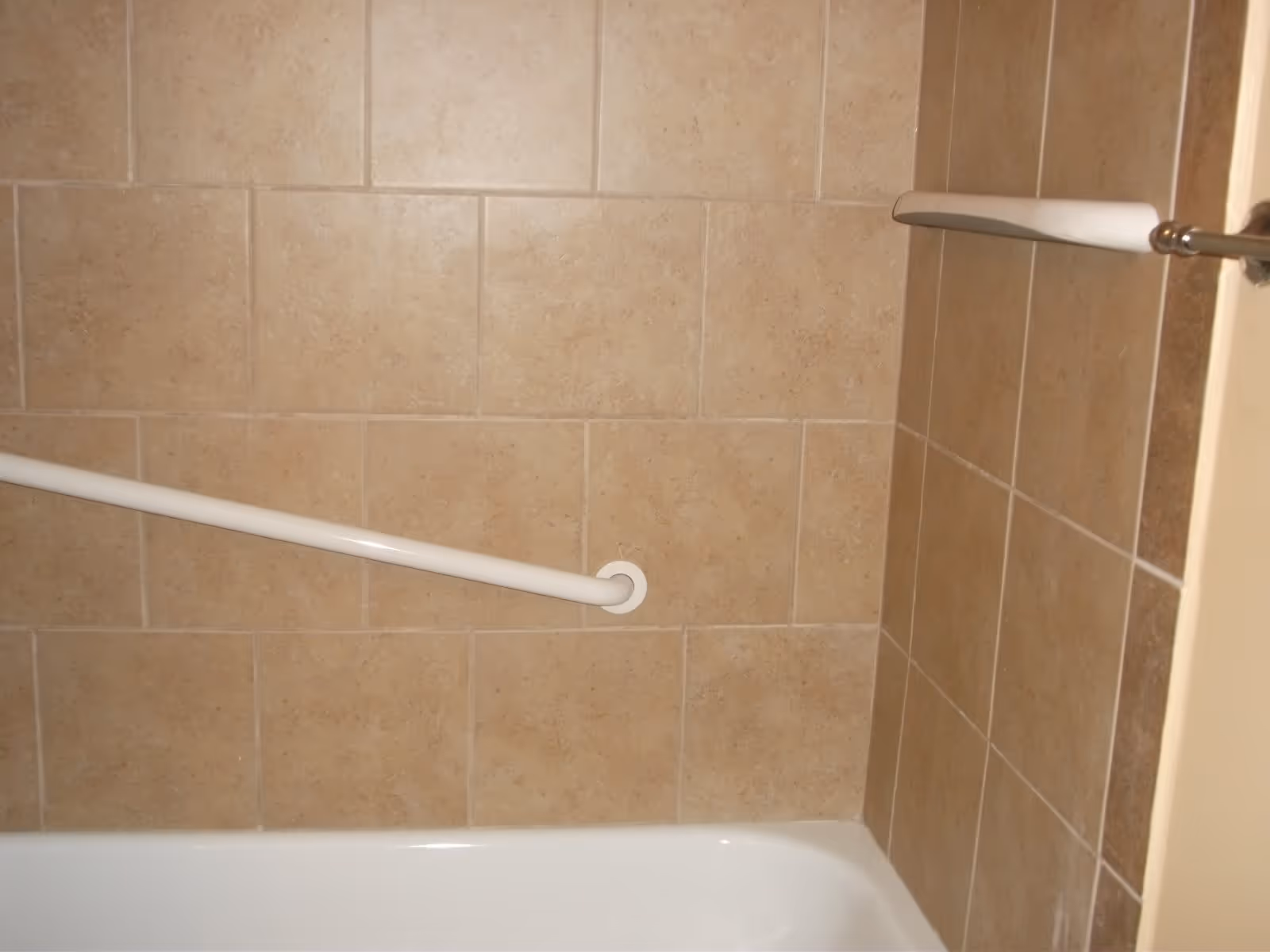 A bathroom shower area with beige tiled walls, a white bathtub, and two white grab bars installed on the walls for support.