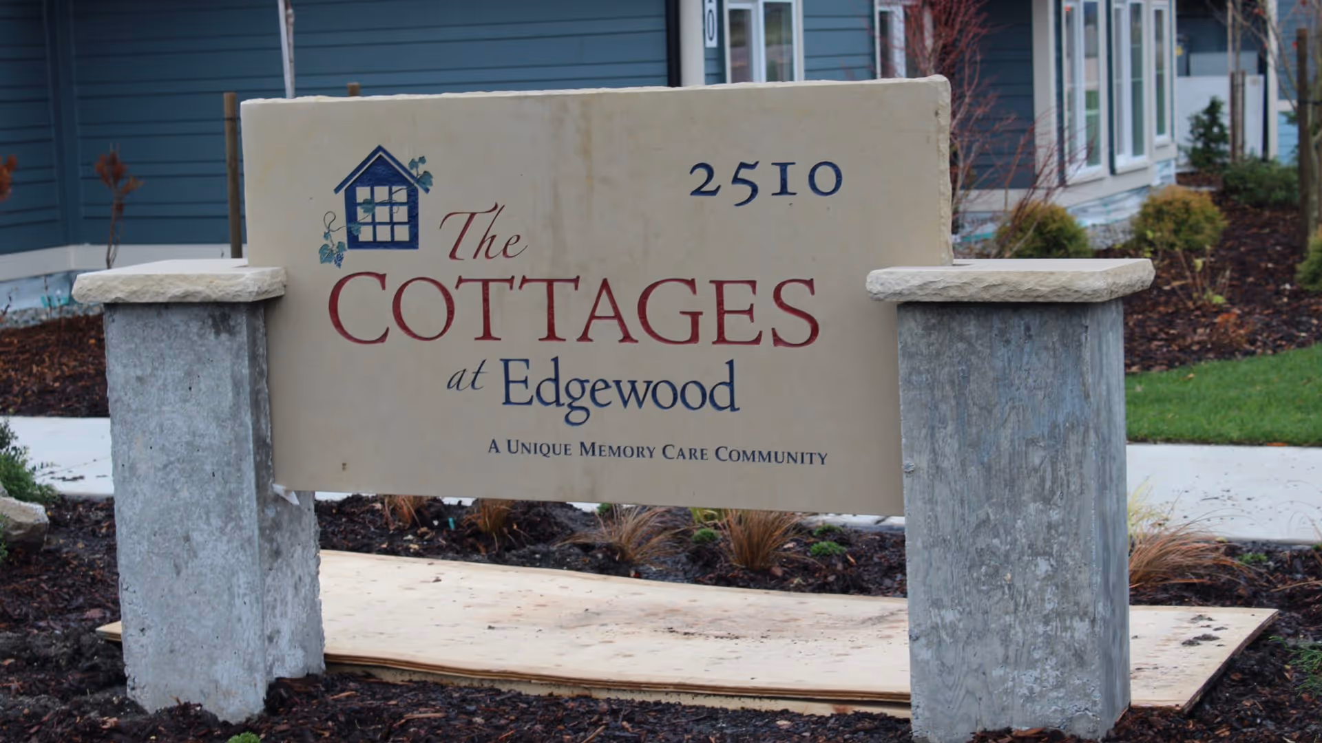 Stone sign for The Cottages at Edgewood, a unique memory care community, with the address number 2510. The sign is supported by two concrete pillars and is situated in a landscaped area with mulch and plants, in front of a blue building.