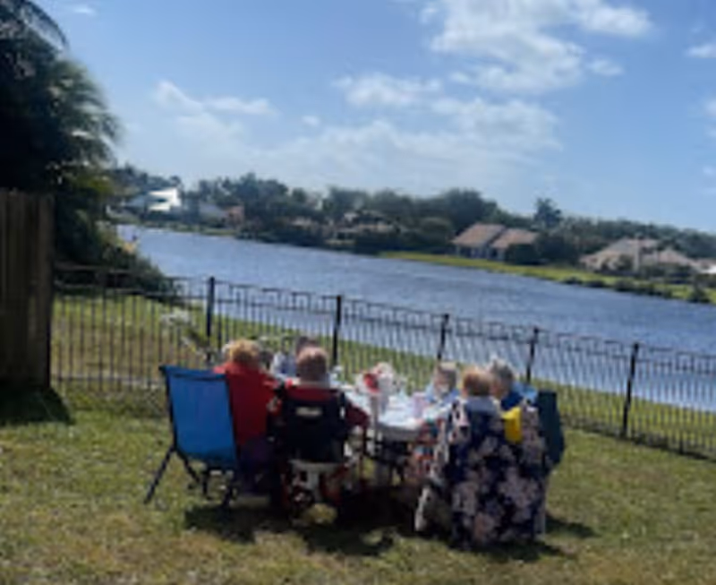 A group of elderly people sitting around a table outdoors near a lake, enjoying a sunny day with a clear blue sky and houses visible across the water.
