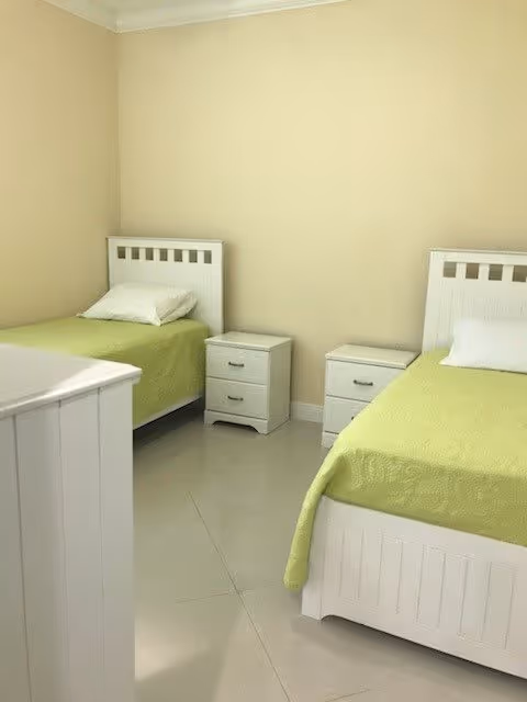 A simple bedroom with two single beds featuring white wooden frames and green bedspreads. Each bed has a white pillow. Between the beds are two small white nightstands with drawers. The walls are beige and the floor is tiled with a light color.