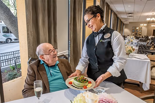 A smiling elderly man seated at a dining table looks up as a server in uniform presents a plate of salad in a dining room.