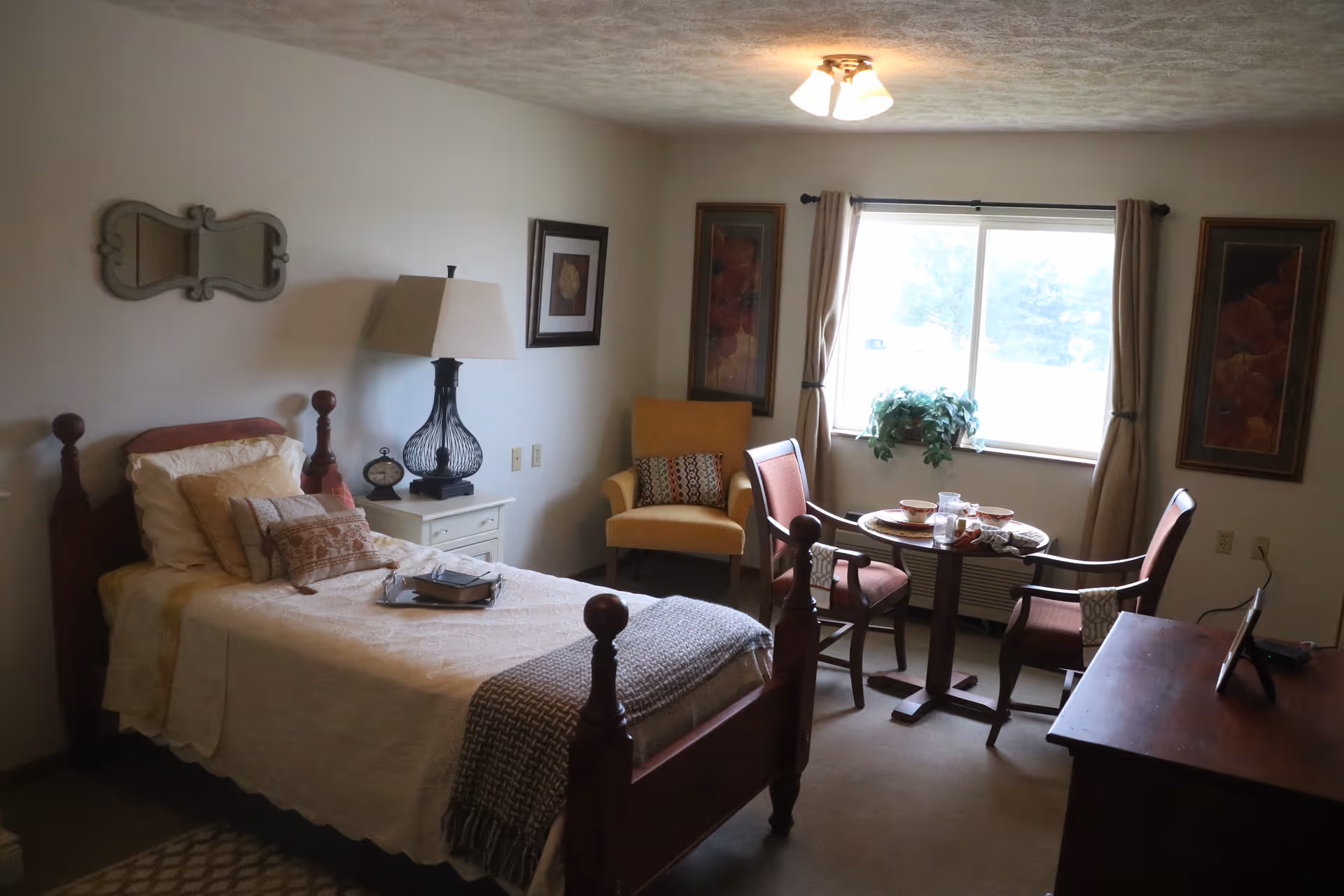 A cozy furnished bedroom with a single bed, nightstand and lamp, and a seating area with chairs and a small table by a window.