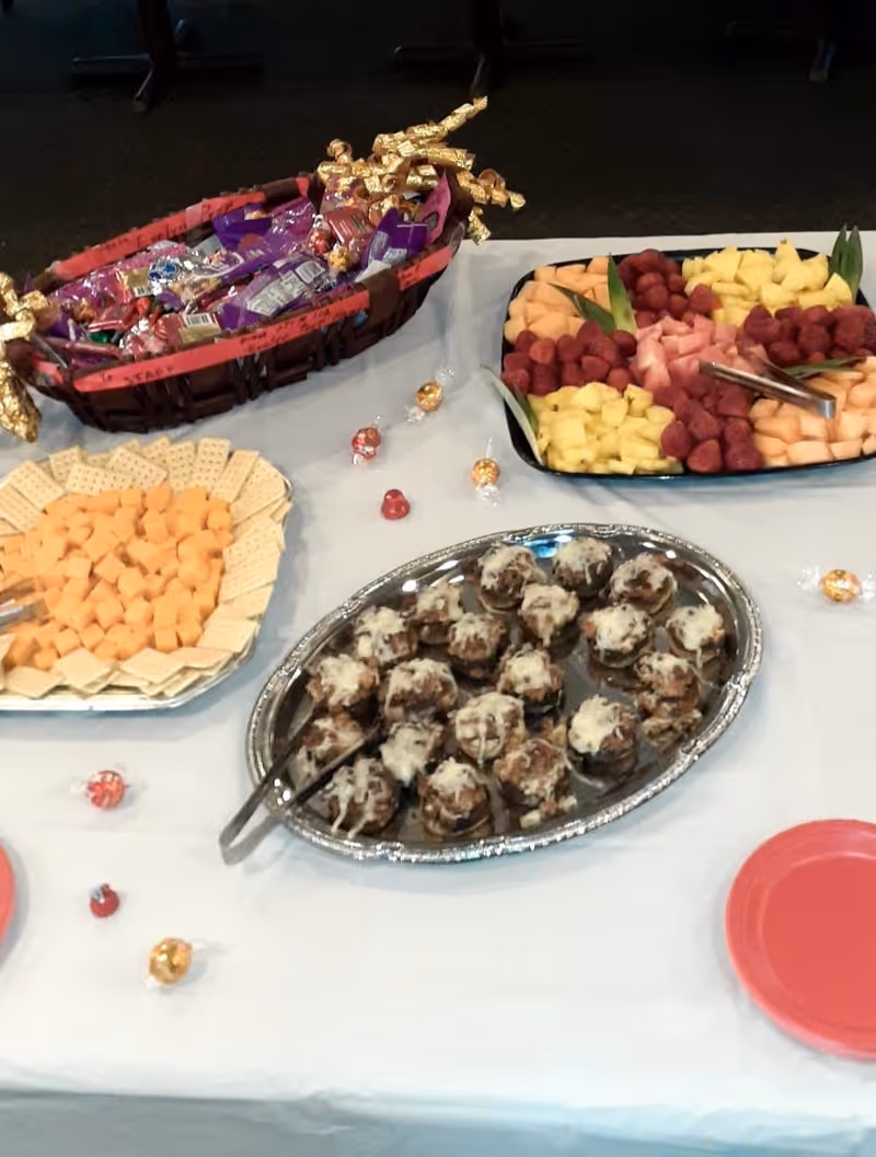 Buffet table with trays of fruit, cheese and crackers, meatball appetizers, and a basket of candy on a white tablecloth.