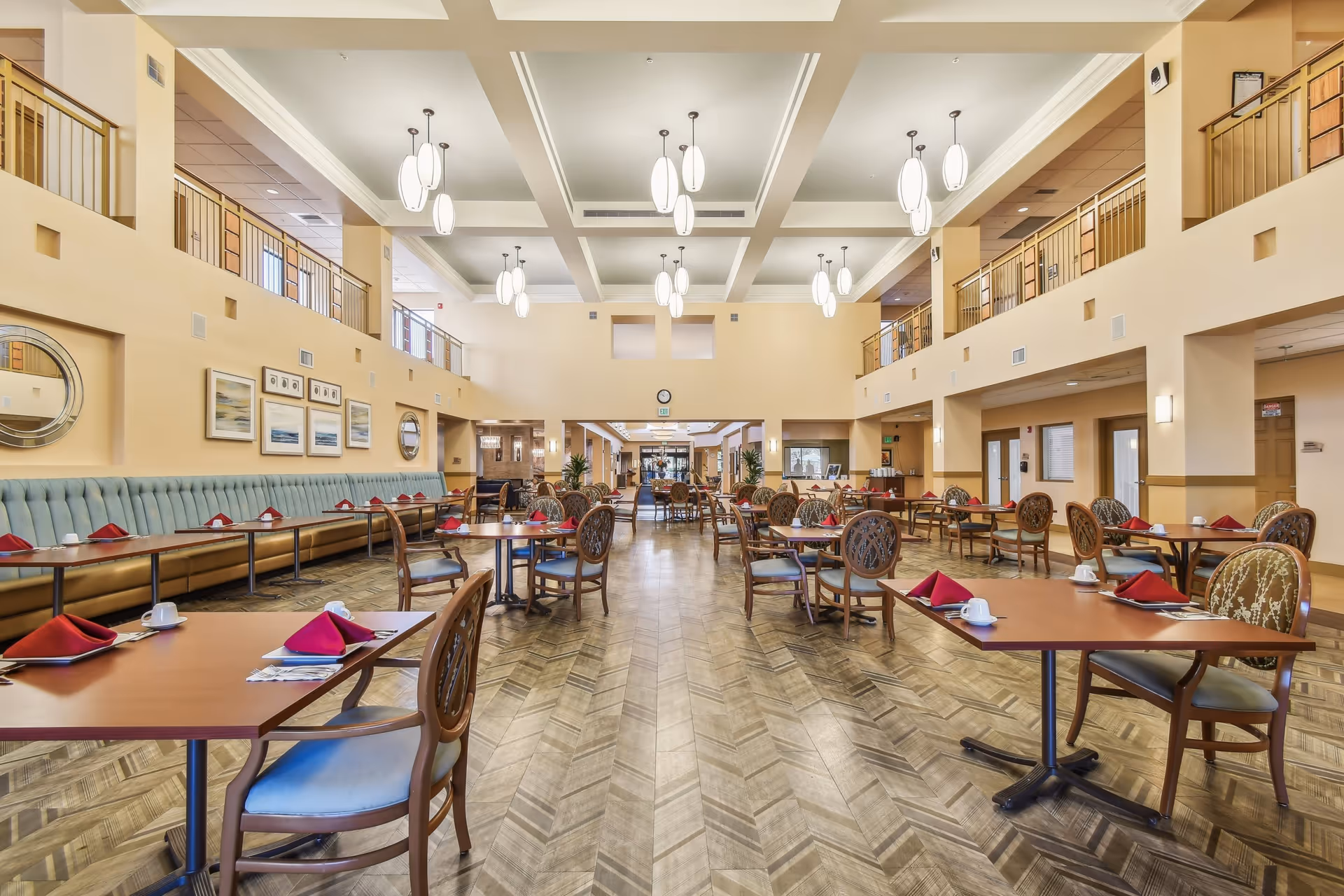 Spacious dining room with multiple wooden tables and chairs arranged neatly. Each table is set with red folded napkins and white cups. The room features high ceilings with modern hanging light fixtures, a long cushioned bench along one wall, framed artwork, and a wooden patterned floor. There is a second-floor balcony railing visible above the dining area.