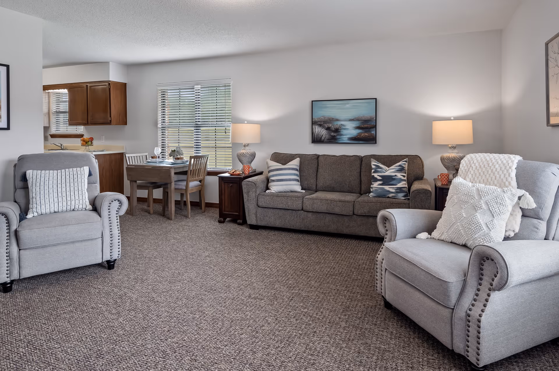 A cozy living room with two gray armchairs, a gray sofa with decorative pillows, two side tables with lamps, a small dining table with four chairs near a window, and a kitchen area with wooden cabinets in the background.