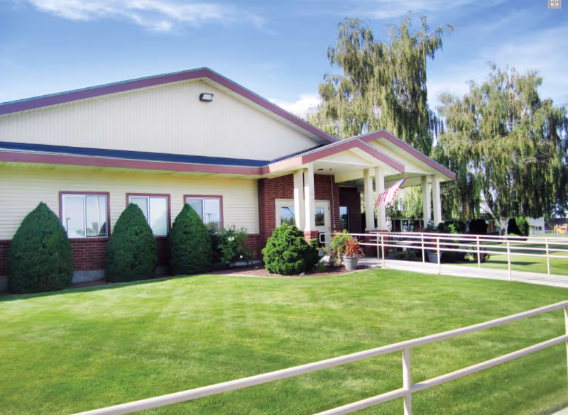 Exterior view of Avalon Health & Rehabilitation Center Pasco showing a single-story building with a covered entrance, an American flag, well-maintained green lawn, shrubs, and trees under a blue sky.
